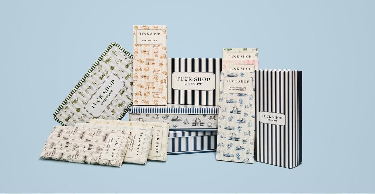 A collection of Tuck Shop chocolate boxes and wrapped chocolates arranged on a light blue background.