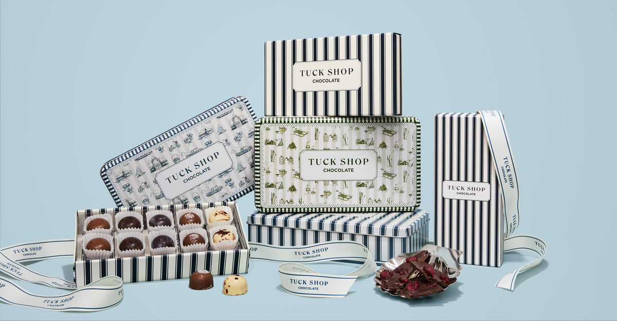 A Tuck Shop chocolate gift set arranged on a light blue background, featuring striped and patterned boxes, an open box of assorted chocolates, and ribbon details.